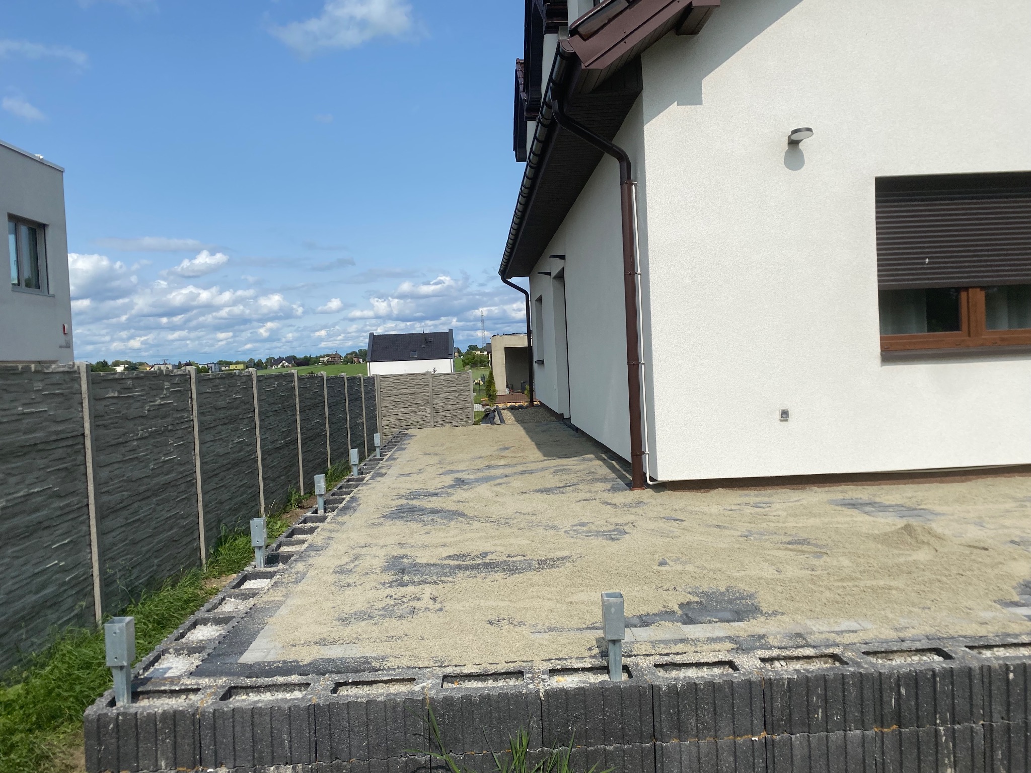 Przygotowany teren pod budowę tarasu przy domu jednorodzinnym. Widoczne bloczki fundamentowe i podsypka piaskowa. W tle ogrodzenie panelowe i błękitne niebo.