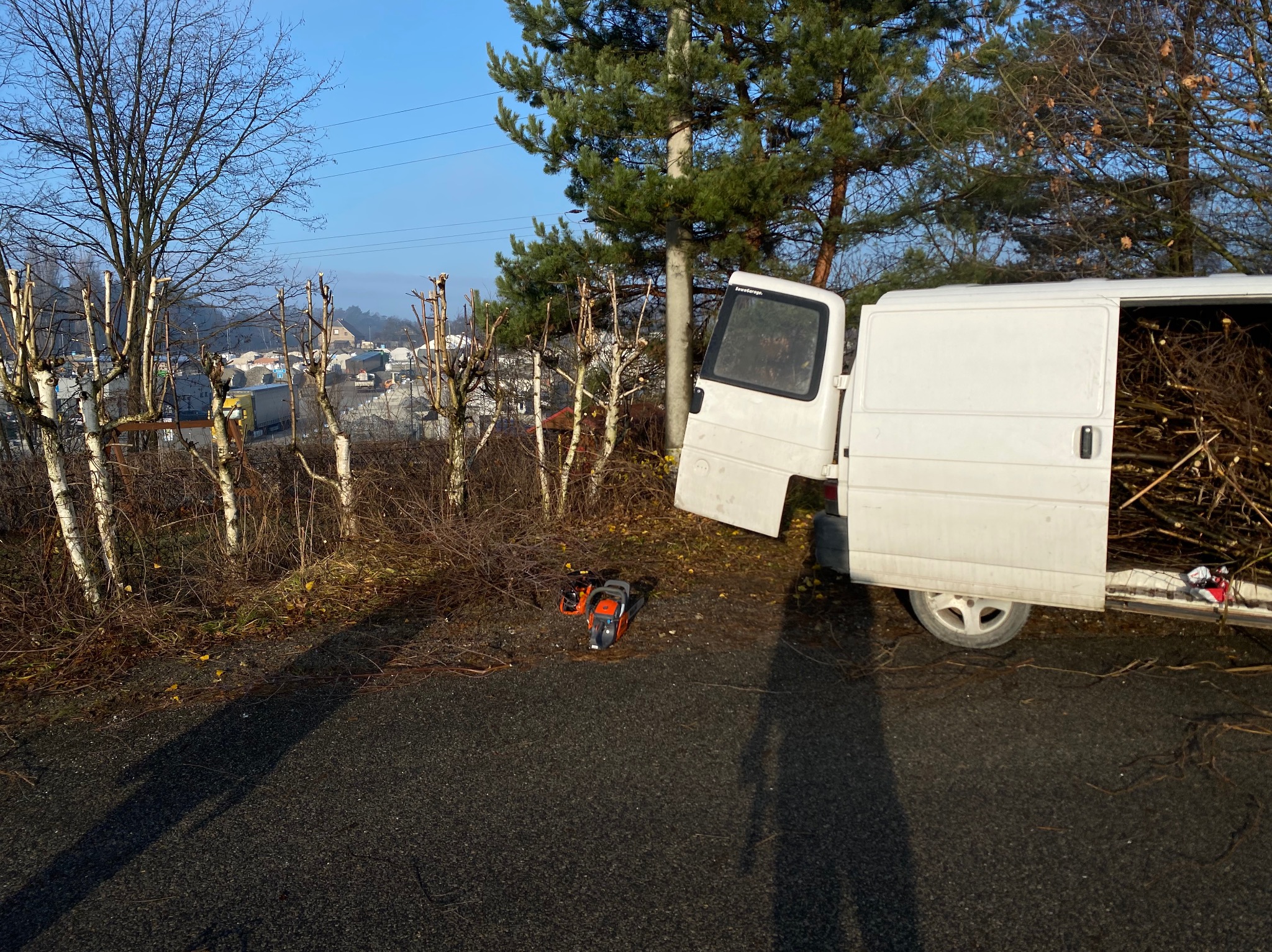 Przycięte drzewa wzdłuż drogi, obok biały bus z otwartymi drzwiami, w środku gałęzie. Na ziemi leży piła spalinowa. Widok na miasto w tle.