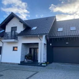 Nowoczesny dom jednorodzinny z czarną dachówką, białymi i grafitowymi elewacjami, ozdobiony świątecznymi dekoracjami. Widoczny balkon z metalową balustradą i brama garażowa.