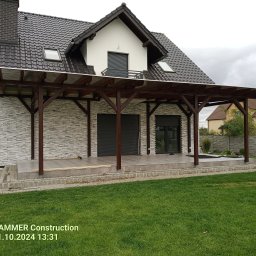 Tynki maszynowe - Wykończony taras z drewnianą pergolą przy domu z elewacją imitującą kamień. Widoczne płytki na tarasie i świeżo położona kostka brukowa. Trawa na pierwszym planie.