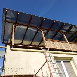 Tynki maszynowe - Zadaszenie balkonu z poliwęglanu na drewnianej konstrukcji, widok z dołu. Ściana budynku z oknami, drabina oparta o ścianę. Błękitne niebo w tle.