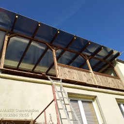 Tynki maszynowe - Zadaszenie balkonu z drewnianą konstrukcją i przezroczystym dachem, widoczne z dołu, z drabiną opartą o ścianę budynku. Błękitne niebo w tle.