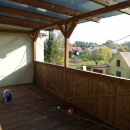 Zadaszony, drewniany balkon z widokiem na okolicę, drewniana podłoga i barierka. Widoczne narzędzia pracy na podłodze balkonu.