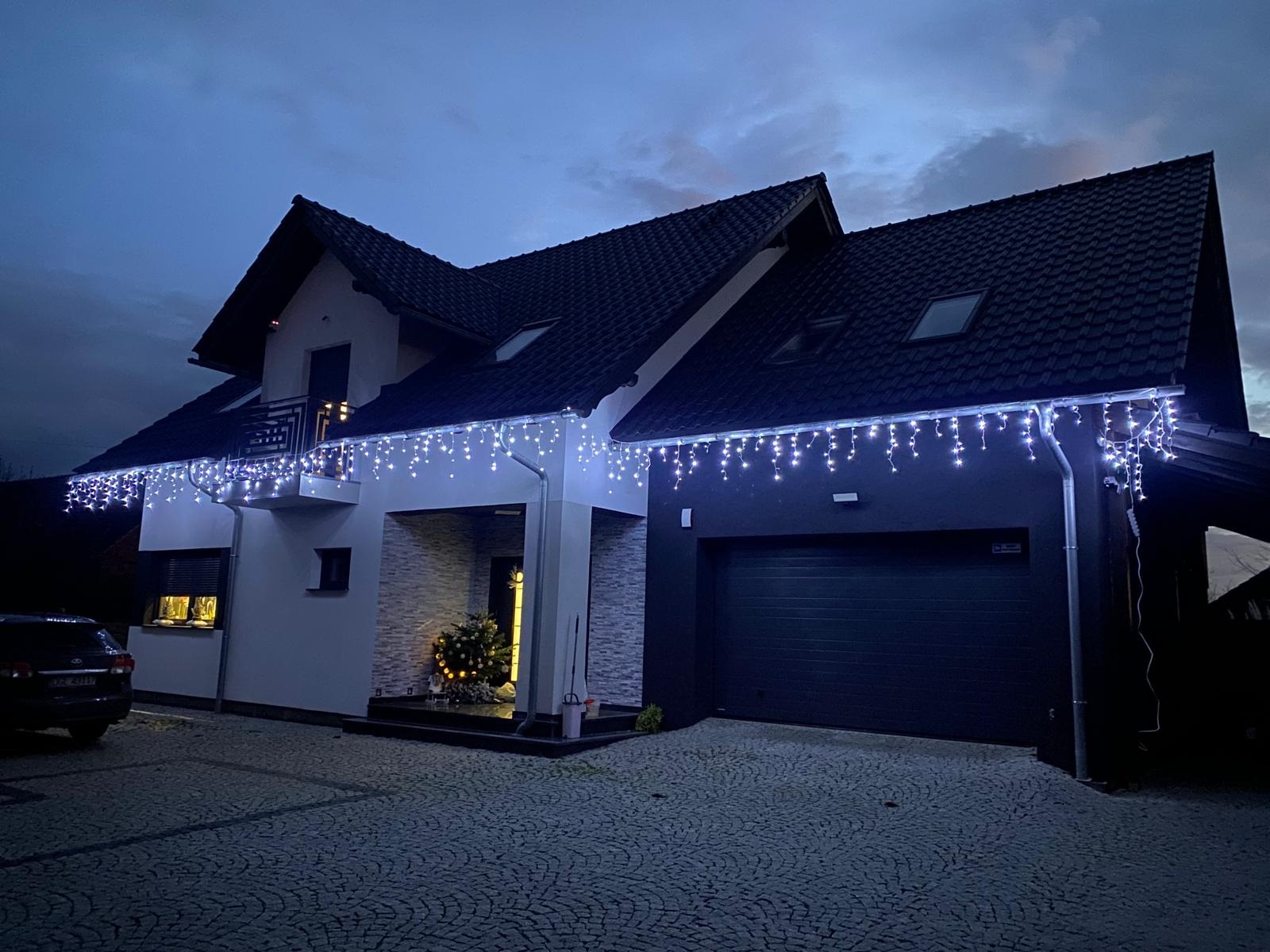 Dwukondygnacyjny dom z ciemnym dachem i dekoracyjnym oświetleniem świątecznym wzdłuż dachu, widoczny samochód i kostka brukowa na podjeździe.