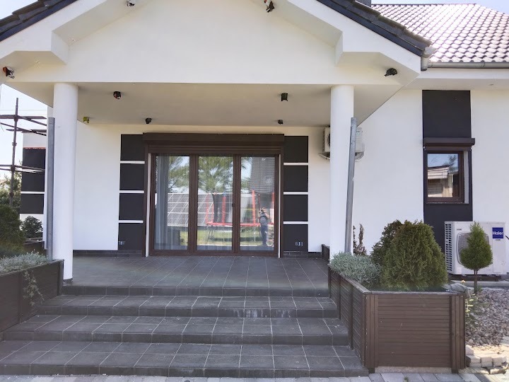 Elewacja domu z ciemnymi akcentami, widoczne kolumny przy wejściu, schody z płytek i ozdobne donice z roślinami. Fragment rusztowania po lewej stronie.