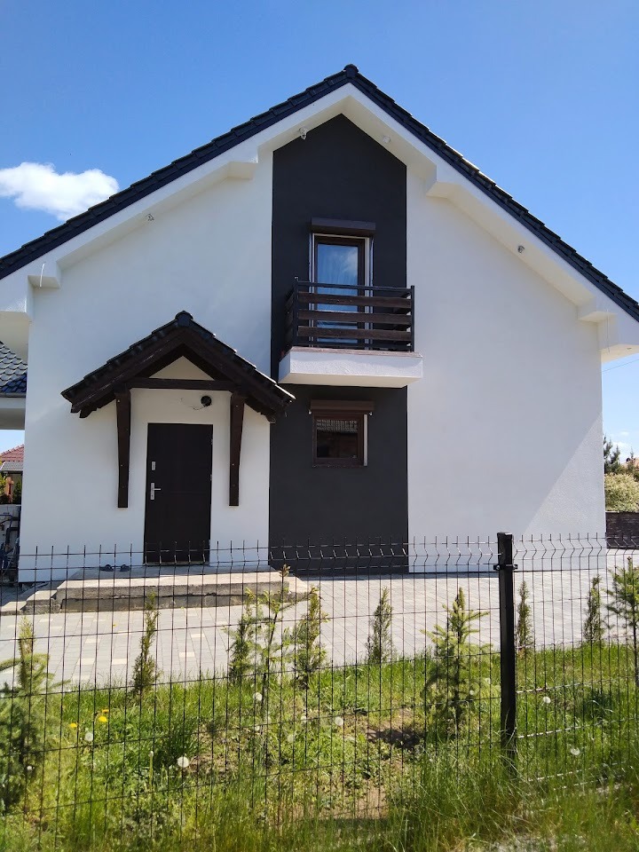 Dwukolorowa elewacja domu jednorodzinnego z balkonem i zadaszonym wejściem, widoczna zza ogrodzenia z młodymi drzewkami iglastymi.