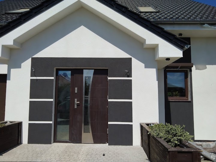 Elewacja domu po remoncie: białe ściany z grafitowymi akcentami wokół drzwi i okna, brązowe drzwi wejściowe i okno z roletą, kostka brukowa, dekoracyjne donice.