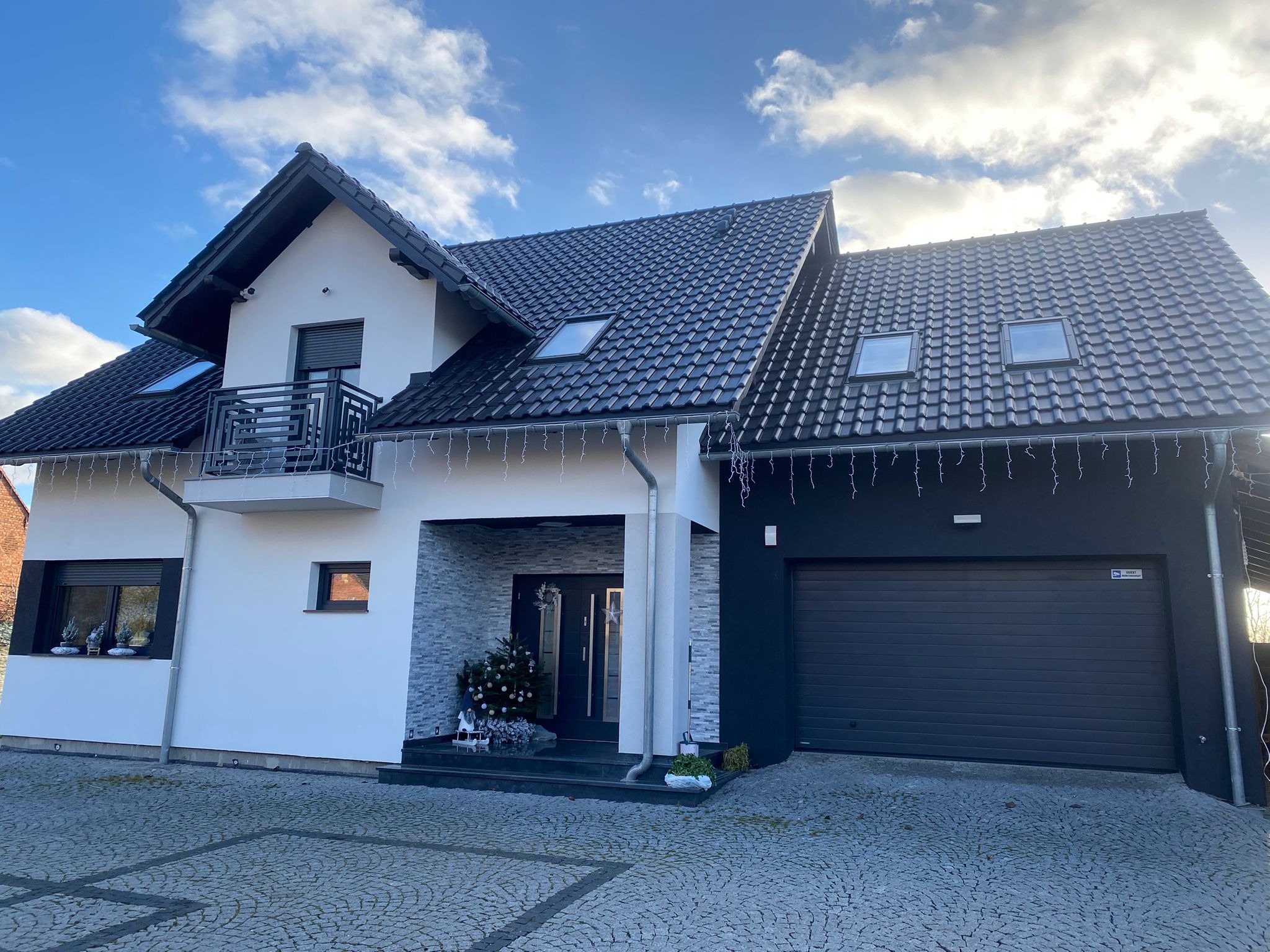 Nowoczesny dom jednorodzinny z czarną dachówką, białymi i grafitowymi elewacjami, ozdobiony świątecznymi dekoracjami. Widoczny balkon z metalową balustradą i brama garażowa.