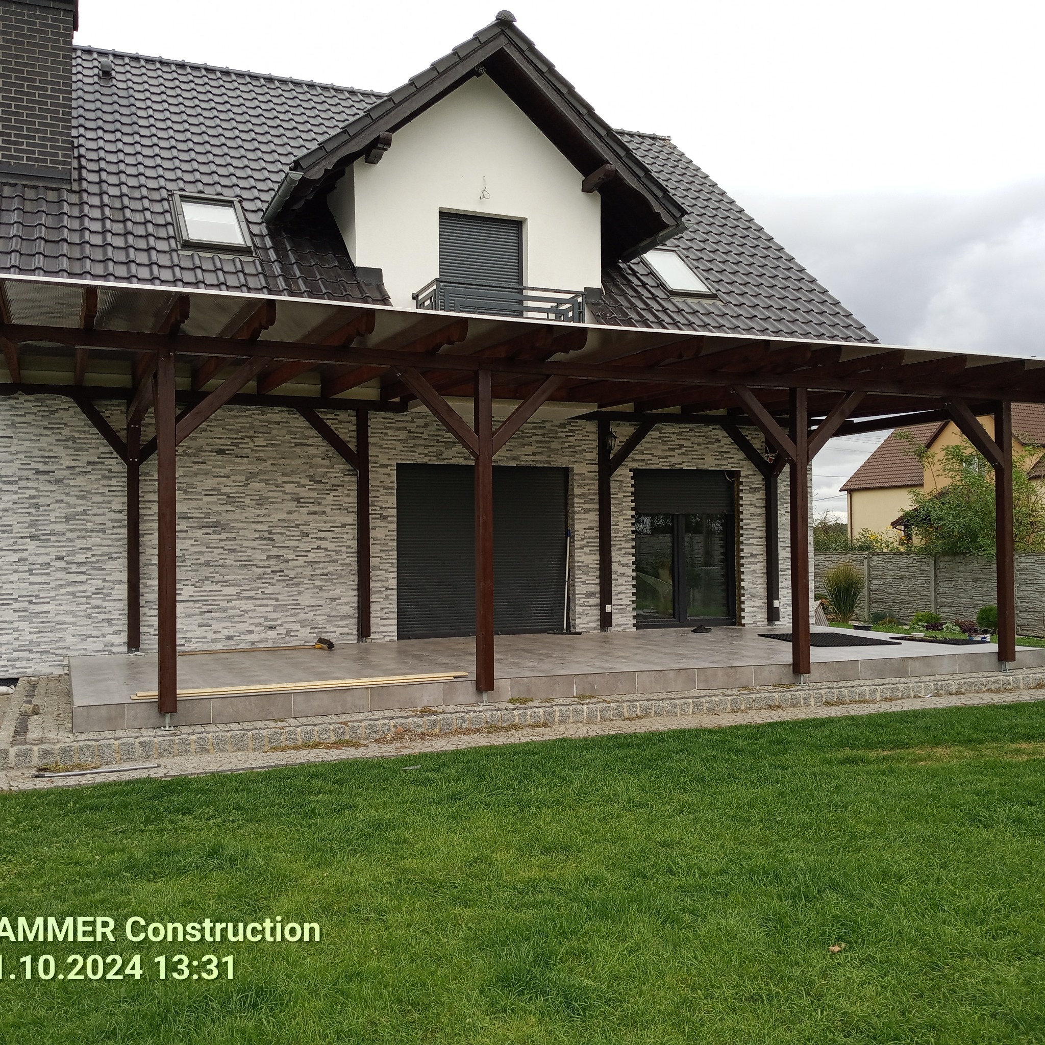 Wykończony taras z drewnianą pergolą przy domu z elewacją imitującą kamień. Widoczne płytki na tarasie i świeżo położona kostka brukowa. Trawa na pierwszym planie.