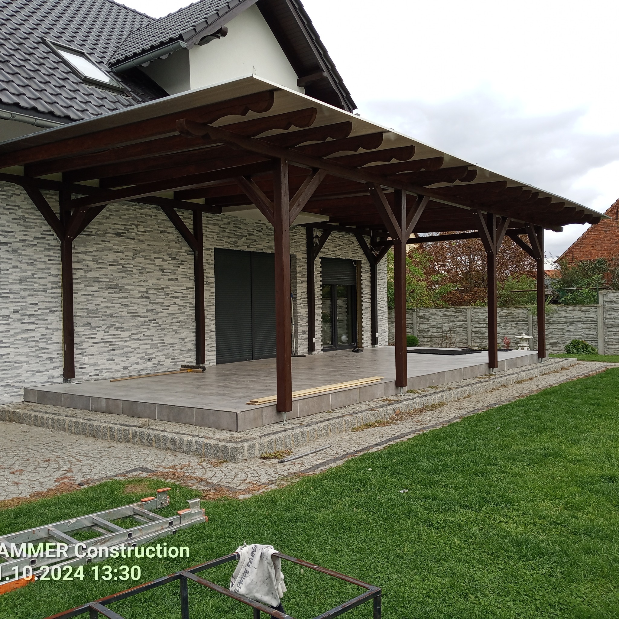 Zadaszenie tarasu z drewnianą konstrukcją i jasną tkaniną, widok z zewnątrz. Na pierwszym planie trawnik i drabina. Tło to elewacja domu.