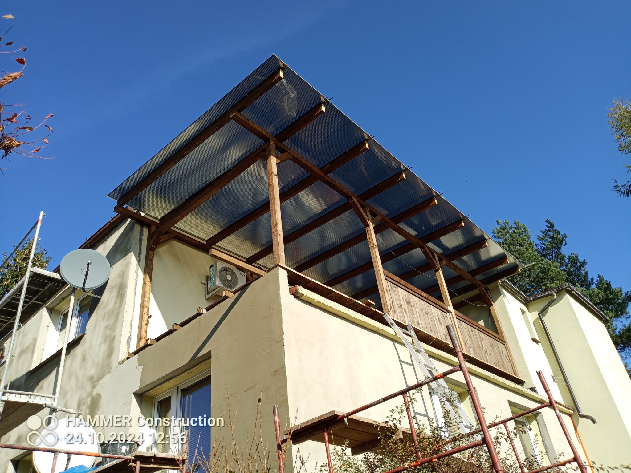 Elewacja budynku w trakcie prac: ocieplenie, tynkowanie. Widoczne rusztowania, częściowo zadaszony balkon z drewnianą balustradą i klimatyzator na ścianie.