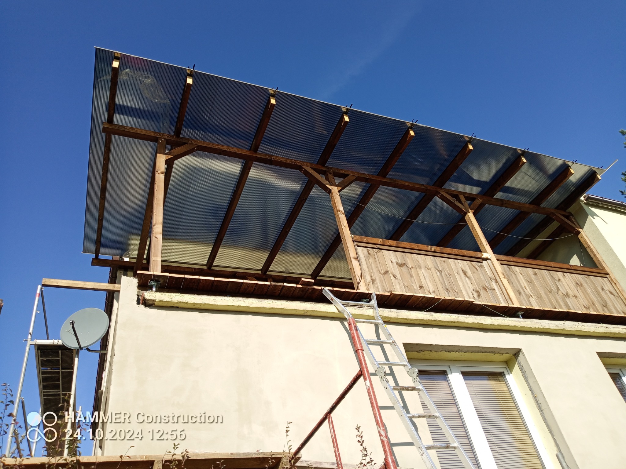 Zadaszenie balkonu z poliwęglanu na drewnianej konstrukcji, widok z dołu. Ściana budynku z oknami, drabina oparta o ścianę. Błękitne niebo w tle.