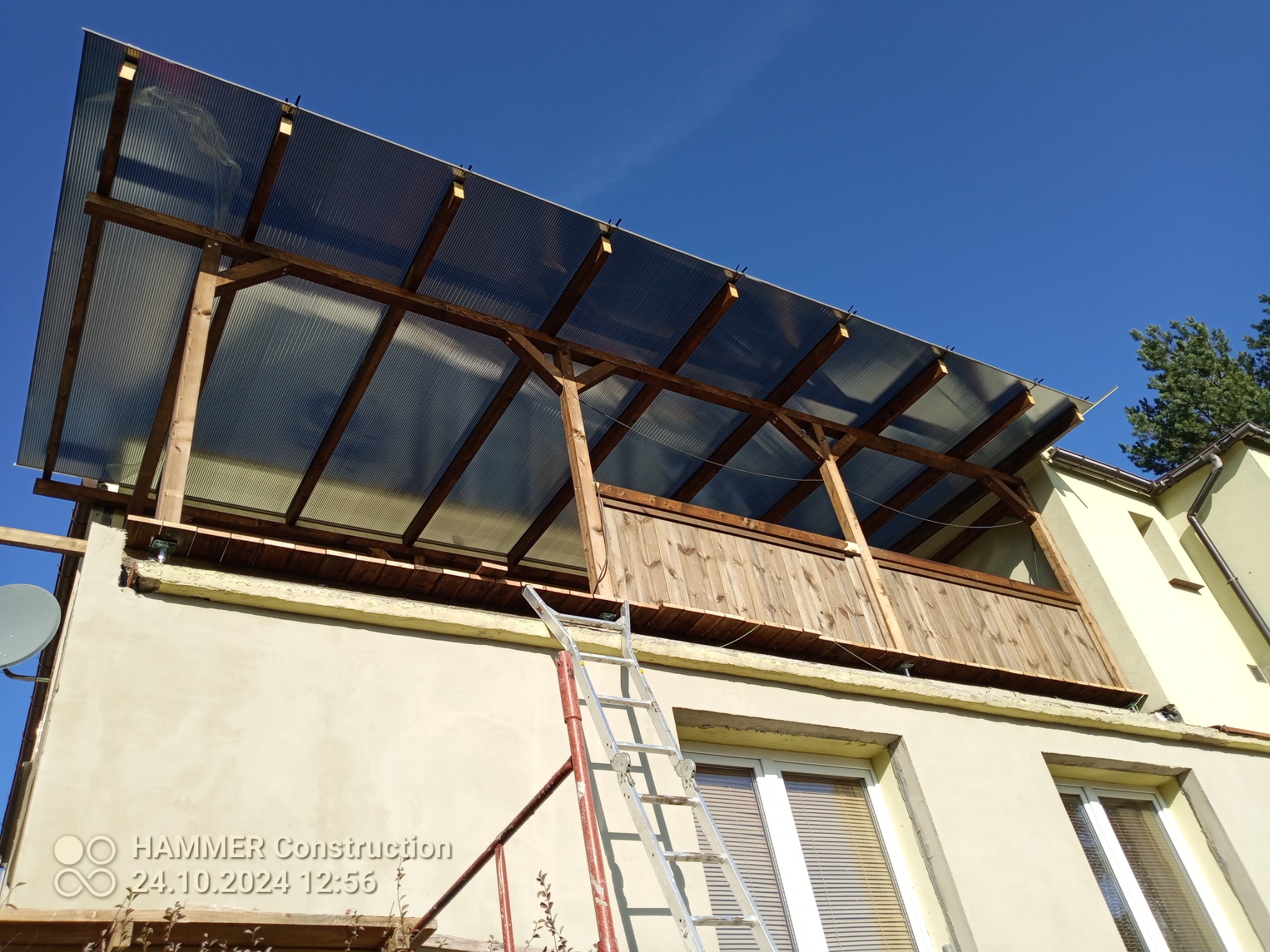 Zadaszenie balkonu z drewnianą konstrukcją i przezroczystym dachem, widoczne z dołu, z drabiną opartą o ścianę budynku. Błękitne niebo w tle.