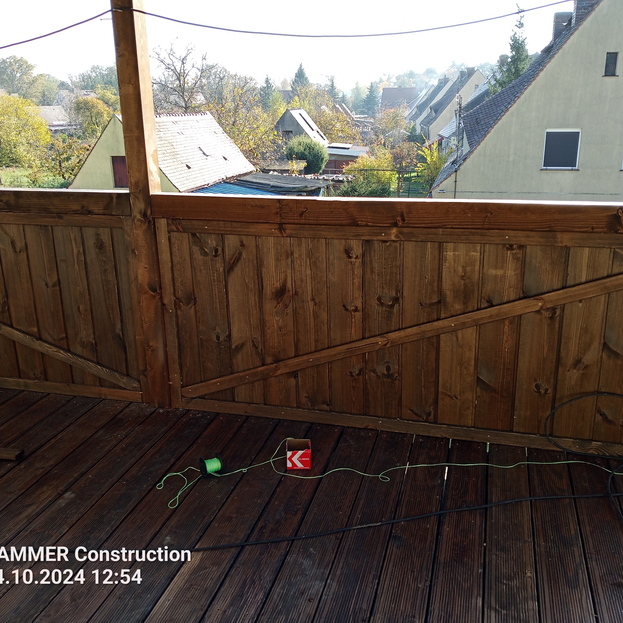Drewniany balkon z widokiem na osiedle w słoneczny dzień. Widoczne narzędzia i materiały budowlane. Deski pomalowane bejcą. Balustrada z desek.