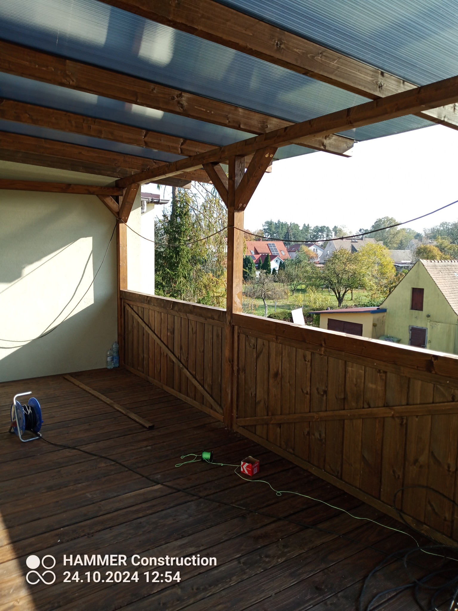 Zadaszony, drewniany balkon z widokiem na okolicę, drewniana podłoga i barierka. Widoczne narzędzia pracy na podłodze balkonu.