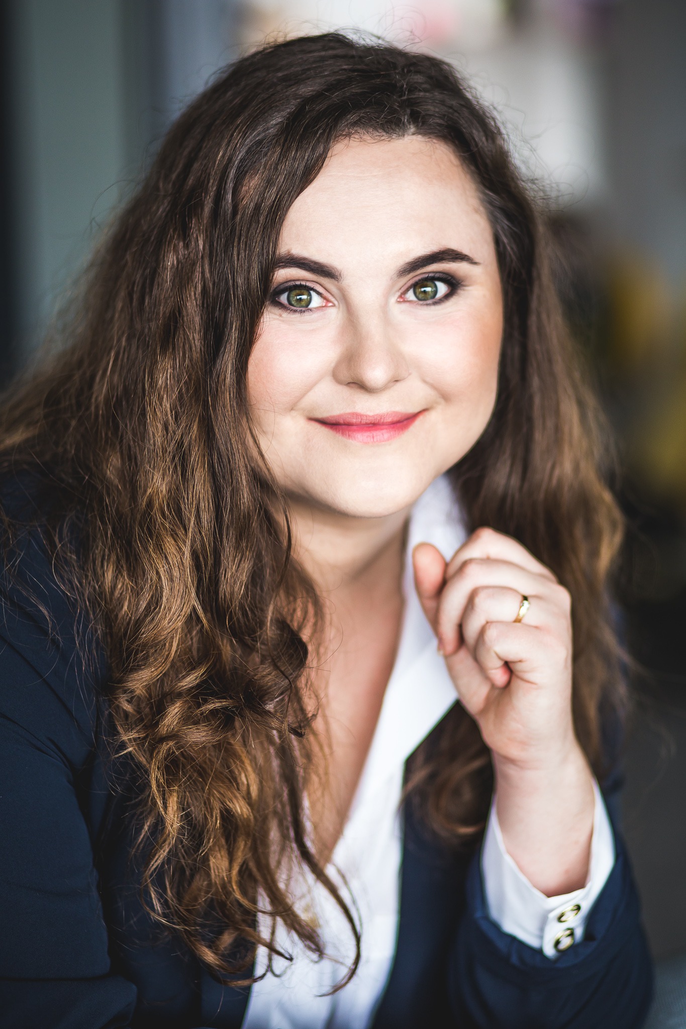 Portret kobiety w ciemnej marynarce i białej koszuli, z delikatnym uśmiechem i pierścionkiem na palcu, na rozmytym tle.