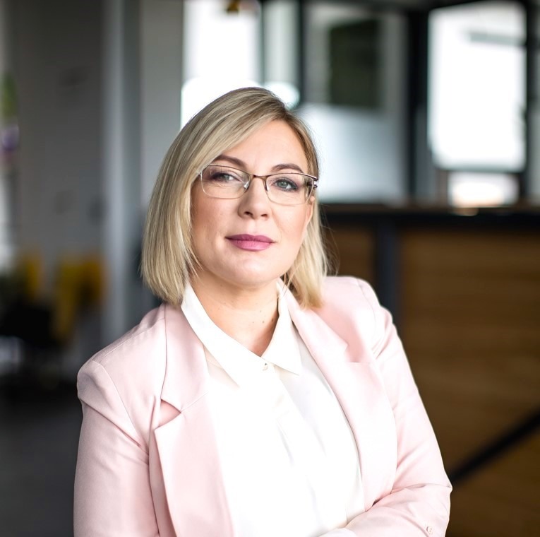 Profesjonalny portret kobiety w okularach i różowym żakiecie, z delikatnym uśmiechem, na rozmytym tle nowoczesnego biura. Naturalne światło.