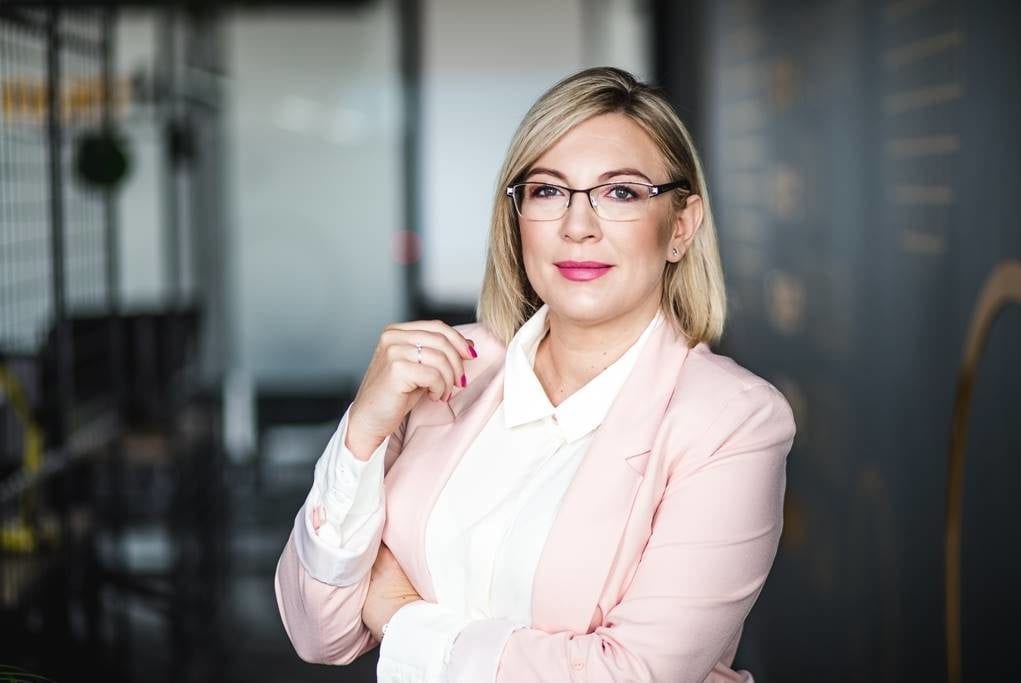 Portret kobiety w okularach i różowym żakiecie, skrzyżowane ręce, stonowane tło biurowe, subtelny uśmiech, profesjonalny wizerunek.