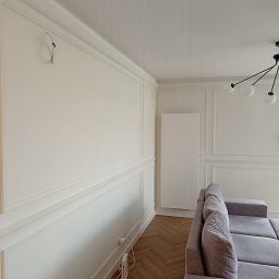 Rem-Rad-Bud Radosław Świętek - Eleganckie wykończenie wnętrza z dekoracyjną sztukaterią ścienną, parkietem w jodełkę i szarą sofą. Widoczne gniazdka i przewody. Minimalistyczna lampa sufitowa.