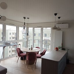 Rem-Rad-Bud Radosław Świętek - Jasne wnętrze: parkiet w jodełkę, okrągły stół z krzesłami w kolorze bordo, wyspa kuchenna z blatem i dekoracyjnymi lamelami. Duże okna z widokiem na miasto.