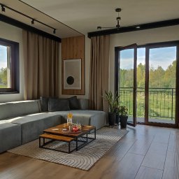 Rem-Rad-Bud Radosław Świętek - Jasne, przestronne wnętrze salonu z szarym narożnikiem, drewnianym stolikiem kawowym i widokiem na zieleń przez balkon. Nowoczesne oświetlenie sufitowe i minimalistyczny design.