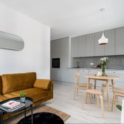 Rem-Rad-Bud Radosław Świętek - Jasne, przestronne wnętrze: złota sofa, okrągły stół i szare szafki kuchenne z ryflowanymi frontami, podkreślone naturalnym światłem. Nowoczesny design.