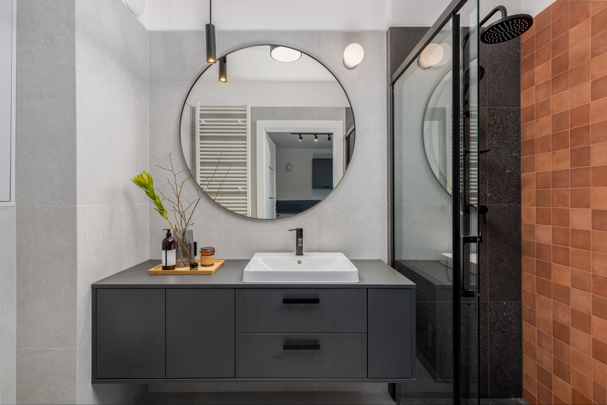 Nowoczesna łazienka z okrągłym lustrem i szarą szafką pod umywalką. Kabina prysznicowa z czarnymi elementami i terakotową ścianą. Minimalistyczny design.