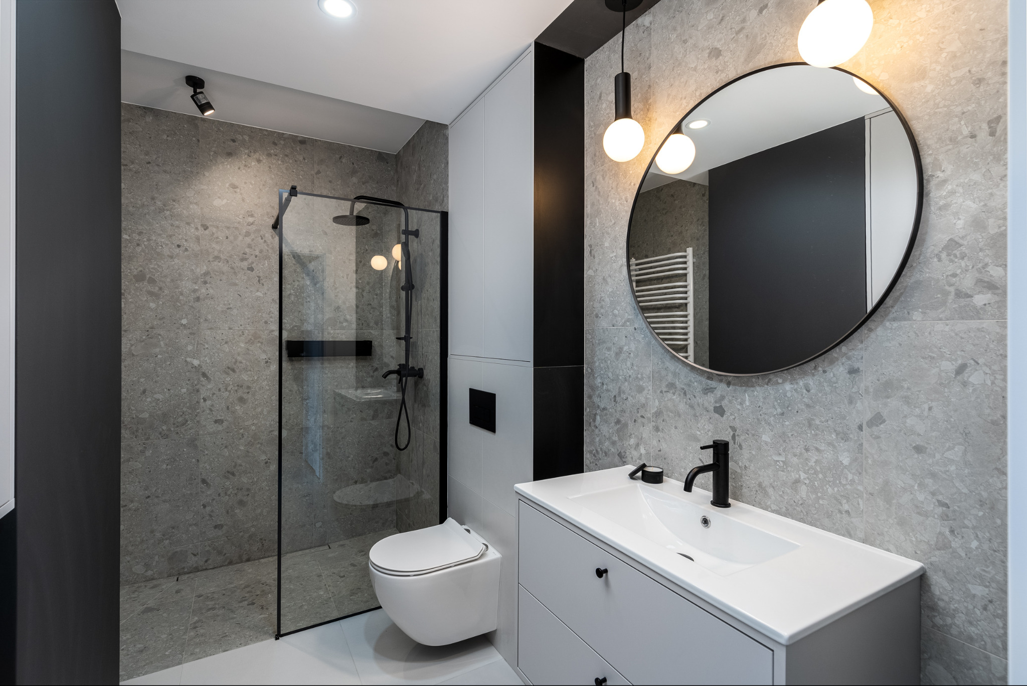 Nowoczesna łazienka w szarościach z kabiną prysznicową, wiszącą toaletą i okrągłym lustrem z oświetleniem. Minimalistyczny design.