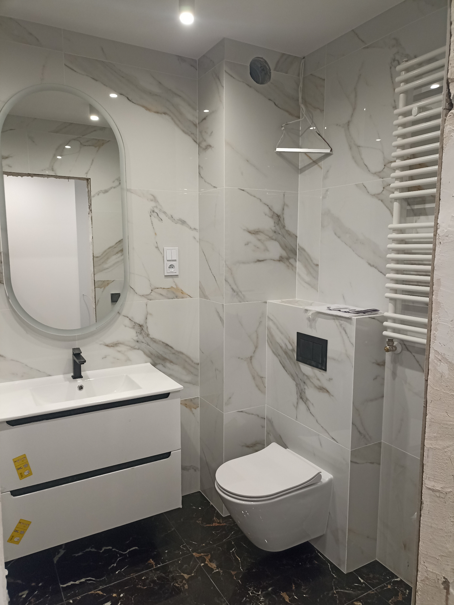 Wykończona łazienka z owalnym lustrem, białą szafką z umywalką, podwieszaną toaletą i grzejnikiem drabinkowym na ścianie z marmurowym wzorem.