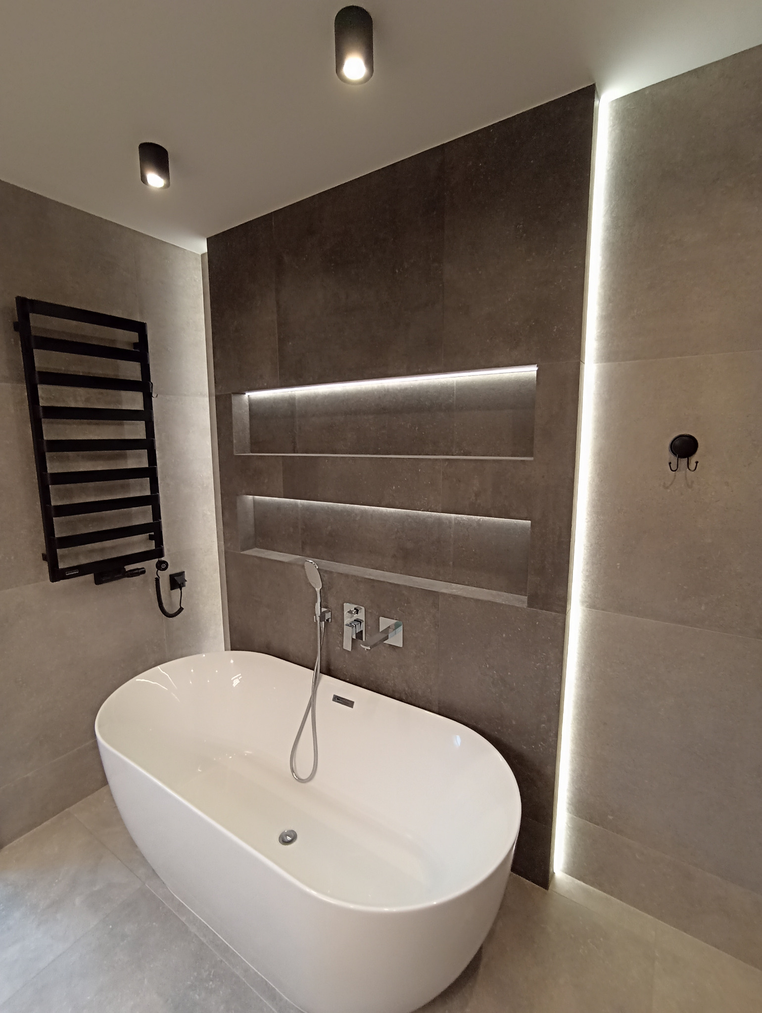 Nowoczesna łazienka z wolnostojącą wanną, grzejnikiem drabinkowym i podświetlanymi wnękami w szarej, kamiennej okładzinie. Minimalistyczny design.