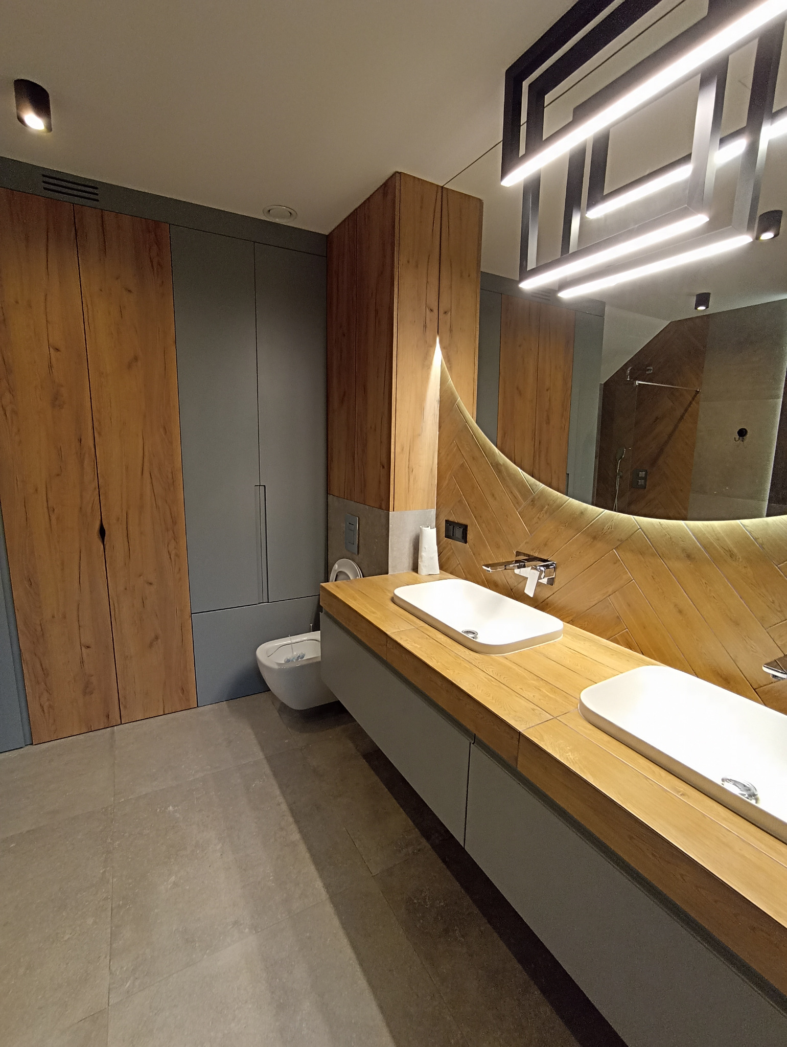 Nowoczesna łazienka z drewnianymi akcentami: szafki z forniru, podwójna umywalka na drewnianym blacie i lustro z podświetleniem LED w kształcie łuku.