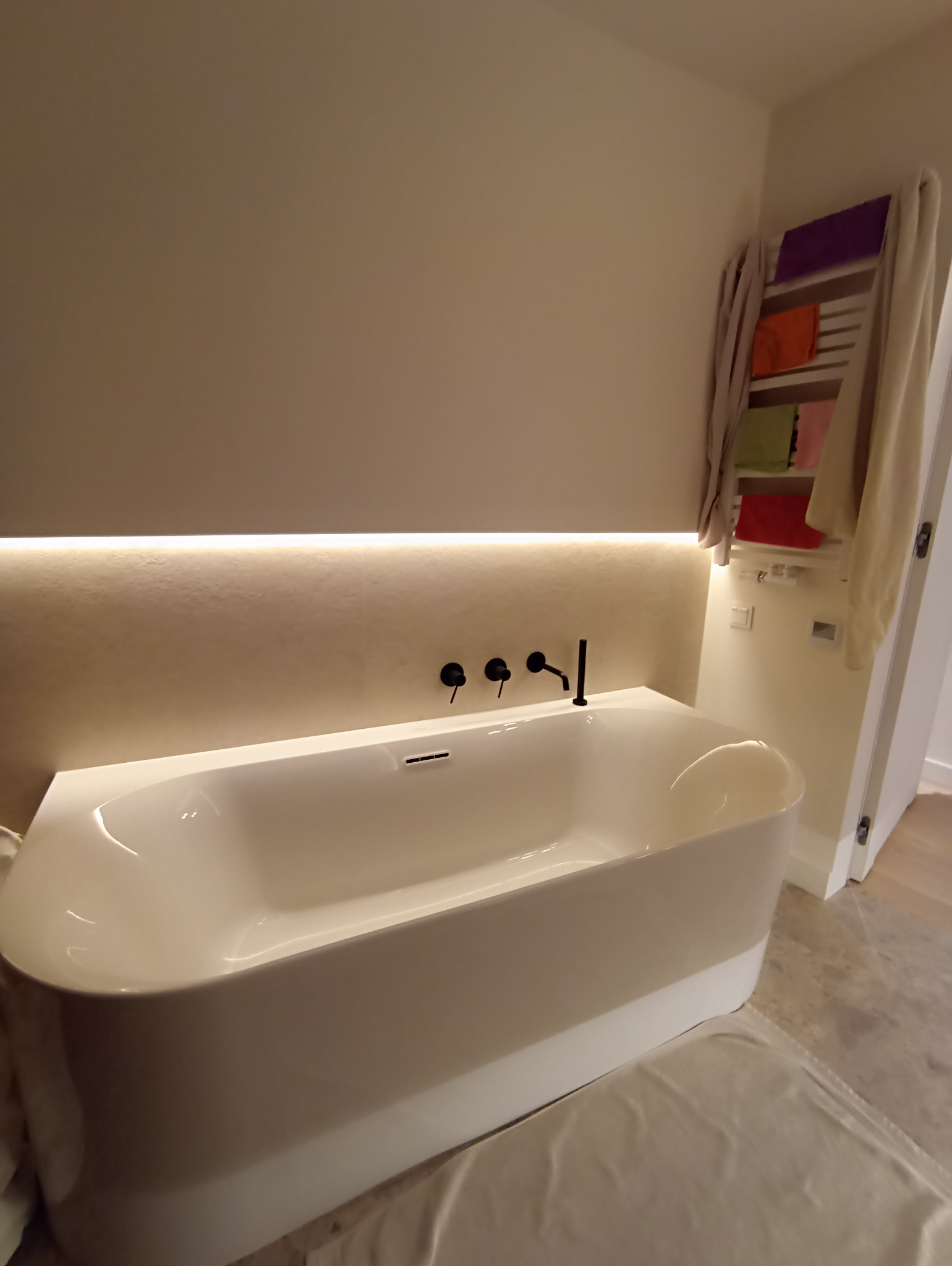 Nowoczesna, wolnostojąca wanna z czarną armaturą na tle podświetlonej ściany i grzejnika drabinkowego z ręcznikami w wykończonej łazience.