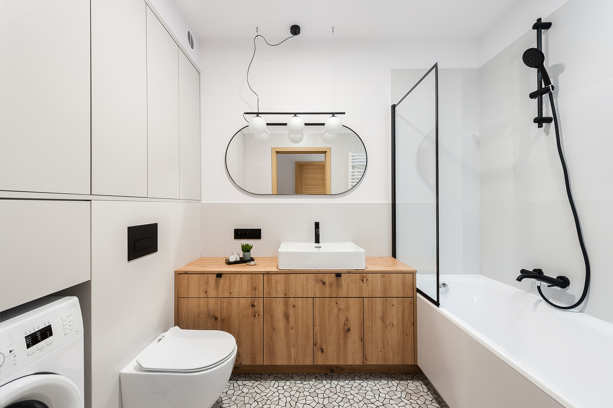 Nowoczesna łazienka z drewnianą szafką pod umywalką, owalnym lustrem i mozaikową podłogą. Uwagę przykuwa czarna armatura i minimalistyczne oświetlenie.