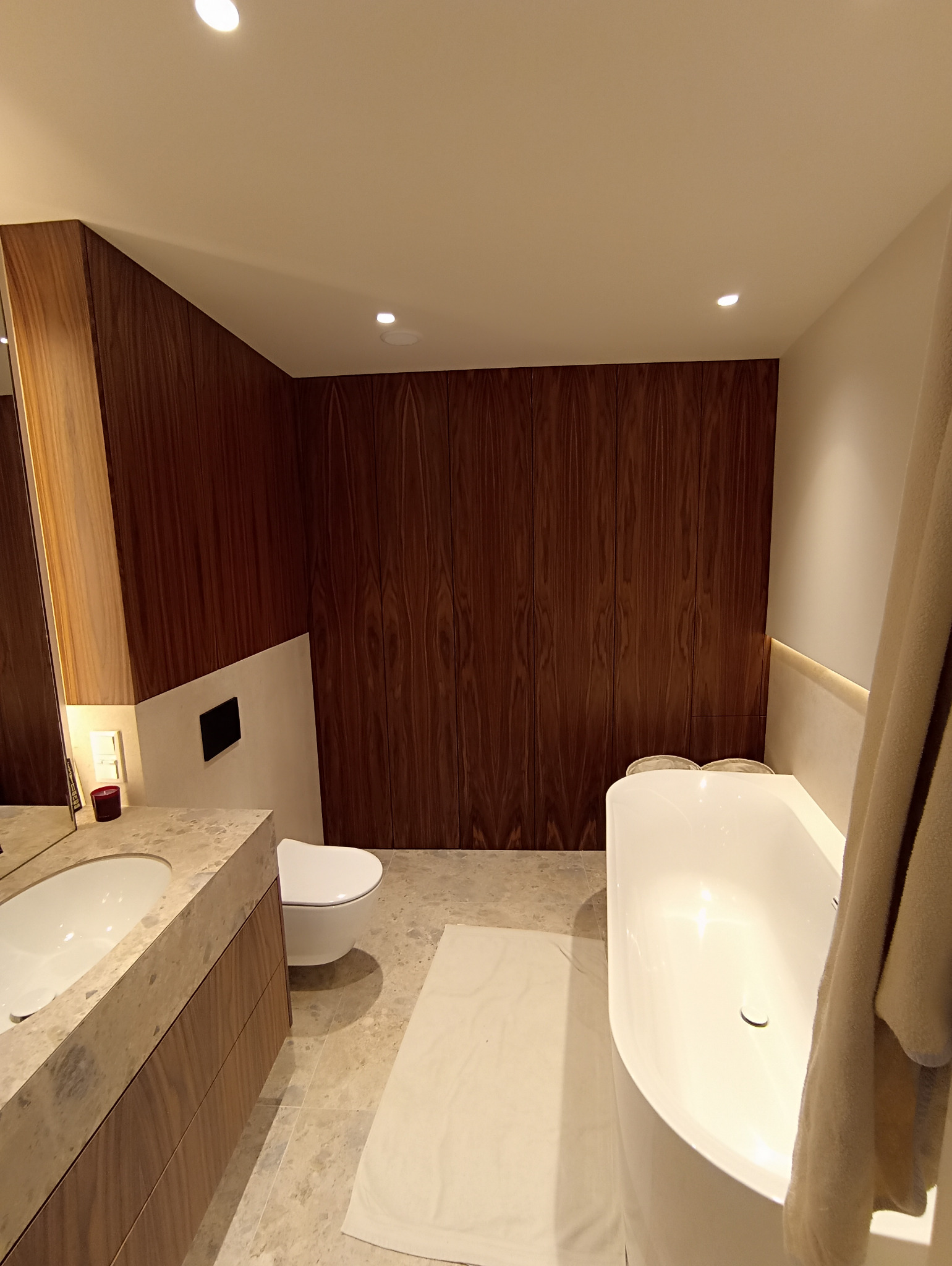 Nowoczesna łazienka z drewnianą zabudową, kamienną umywalką i białą wanną. Minimalistyczny design, ciepłe oświetlenie, beżowe płytki podłogowe.