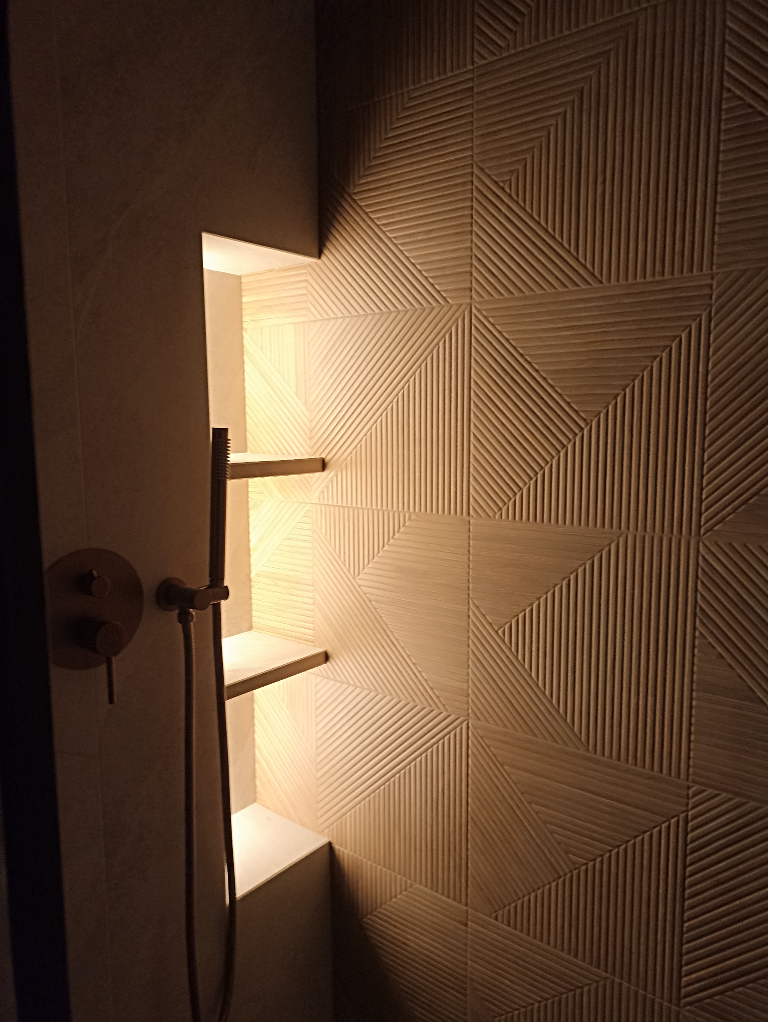 Nowoczesna łazienka z geometryczną ścianą z płytek, wnęką z półkami i złotą armaturą prysznicową. Ciepłe oświetlenie tworzy przytulną atmosferę.