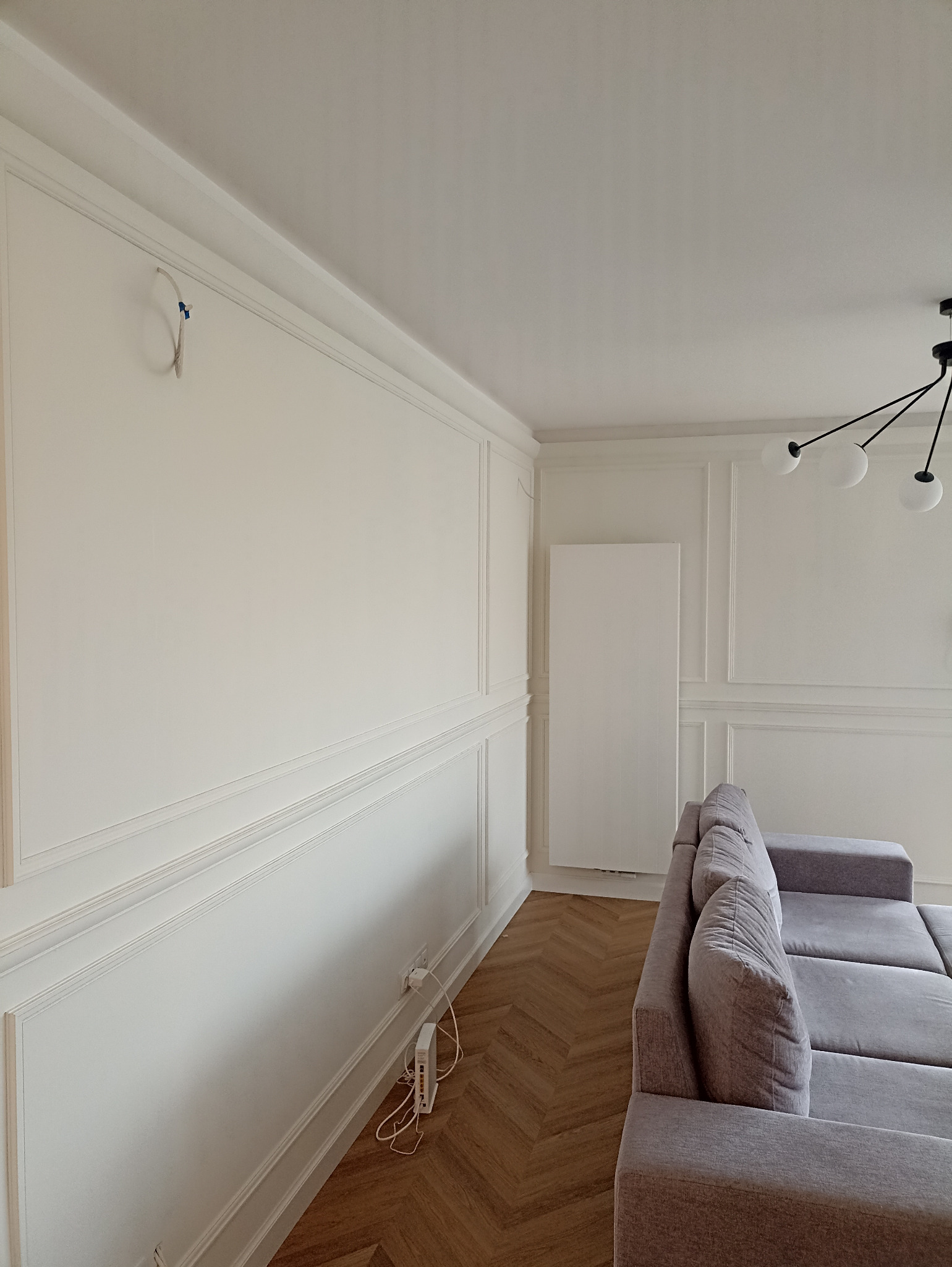 Eleganckie wykończenie wnętrza z dekoracyjną sztukaterią ścienną, parkietem w jodełkę i szarą sofą. Widoczne gniazdka i przewody. Minimalistyczna lampa sufitowa.