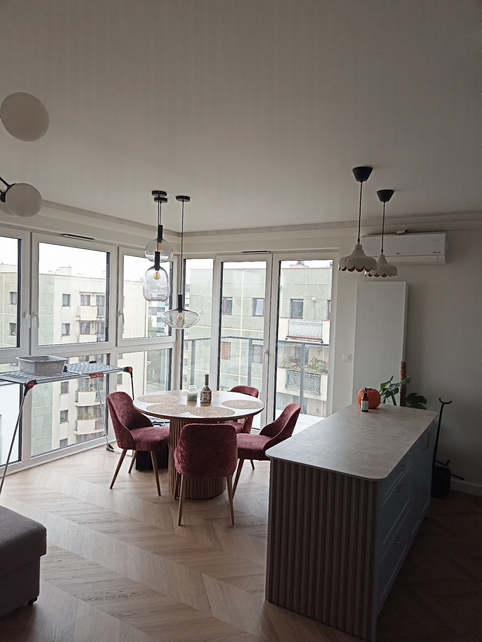 Jasne wnętrze: parkiet w jodełkę, okrągły stół z krzesłami w kolorze bordo, wyspa kuchenna z blatem i dekoracyjnymi lamelami. Duże okna z widokiem na miasto.