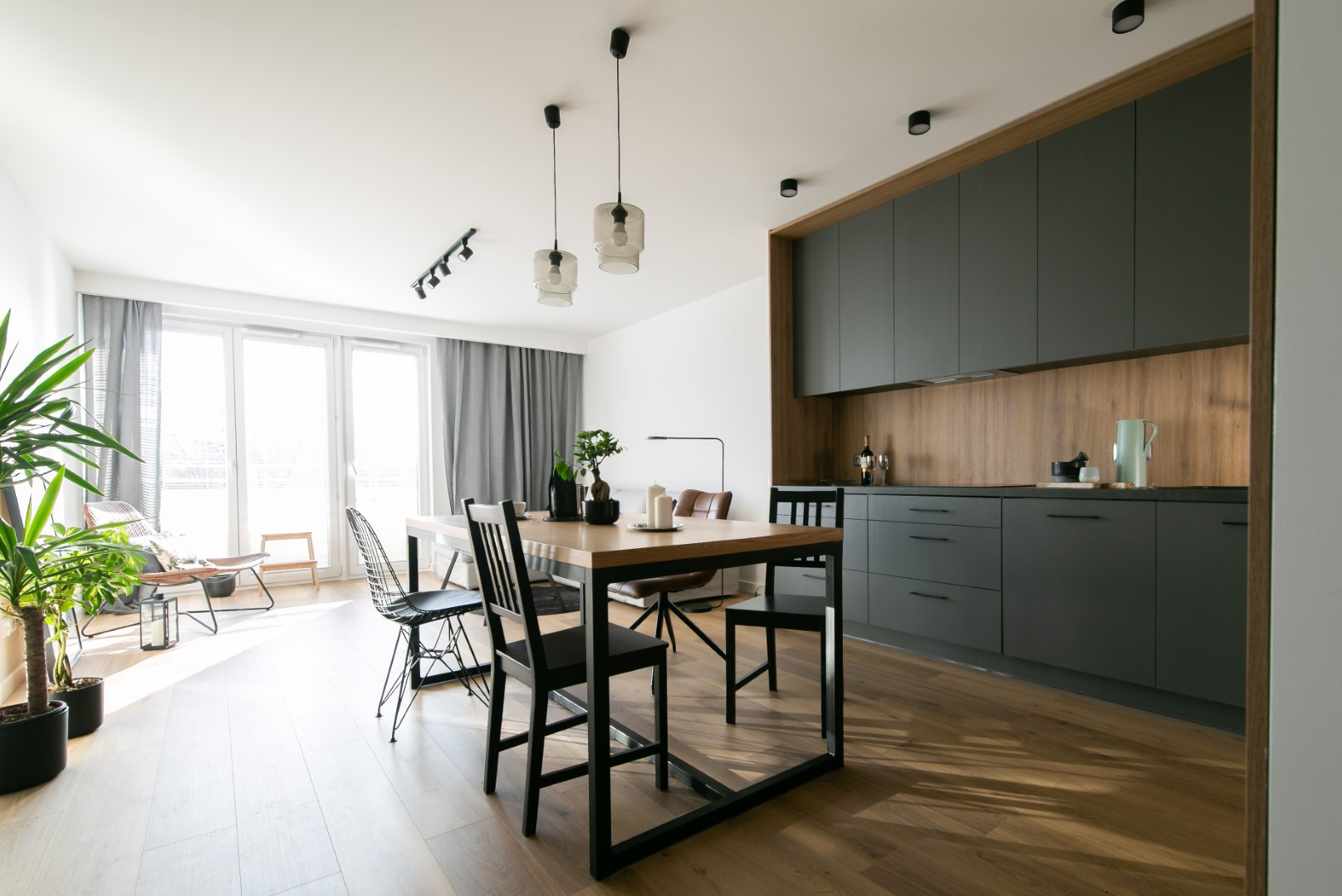 Nowoczesna kuchnia z szarymi szafkami, drewnianym blatem i stołem z czarnymi krzesłami. Jasne wnętrze z dużym oknem i roślinami doniczkowymi.