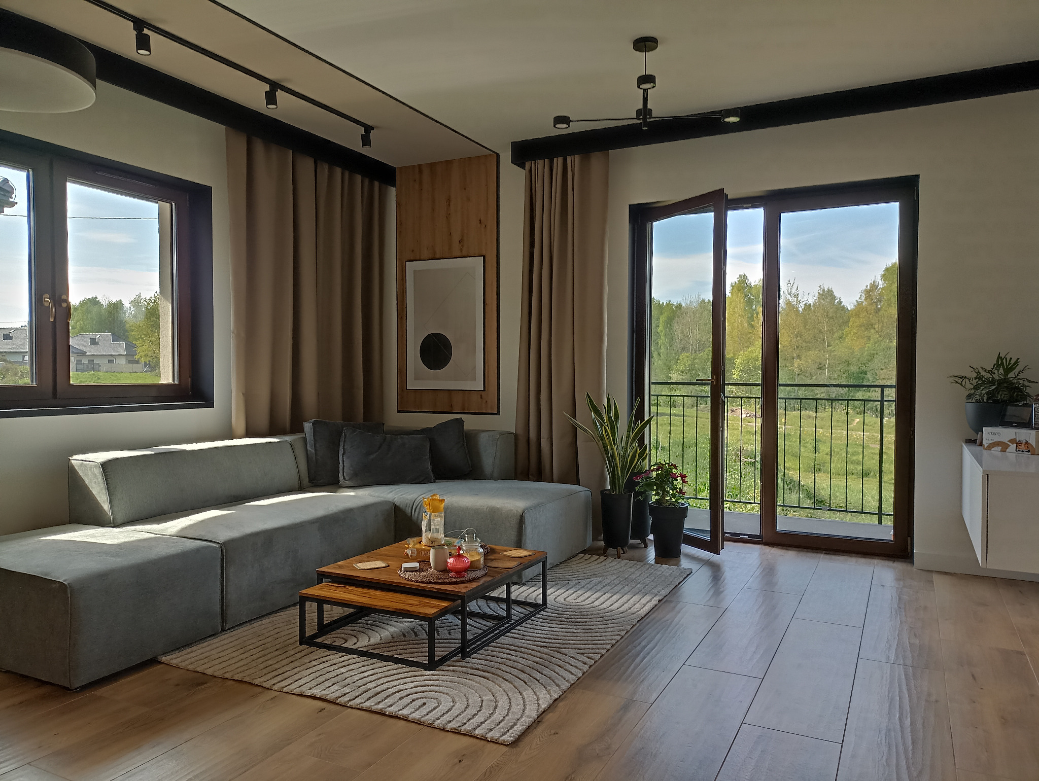 Jasne, przestronne wnętrze salonu z szarym narożnikiem, drewnianym stolikiem kawowym i widokiem na zieleń przez balkon. Nowoczesne oświetlenie sufitowe i minimalistyczny design.