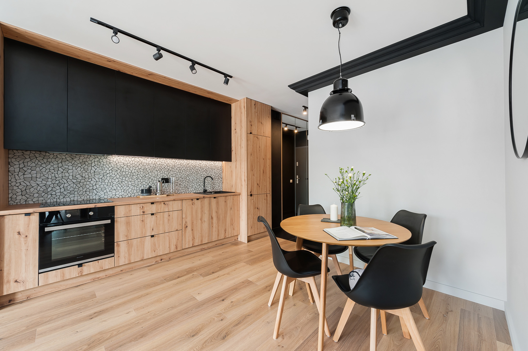 Nowoczesna kuchnia z drewnianymi szafkami i czarnymi akcentami, oświetlona lampą wiszącą nad okrągłym stołem. Na blacie mozaika z kamienia.