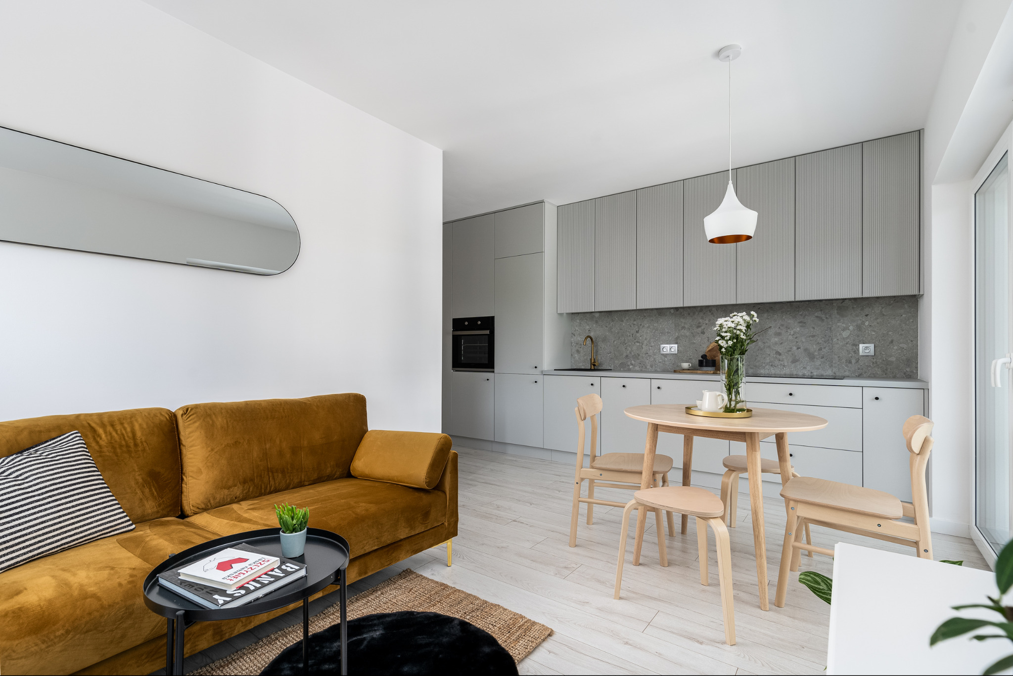 Jasne, przestronne wnętrze: złota sofa, okrągły stół i szare szafki kuchenne z ryflowanymi frontami, podkreślone naturalnym światłem. Nowoczesny design.