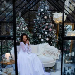 Sesja w szklarni w Dębicy: Uśmiechnięta kobieta w białej sukni siedzi na sofie wśród świątecznych dekoracji, choinek i prezentów. Zimowa sceneria.
