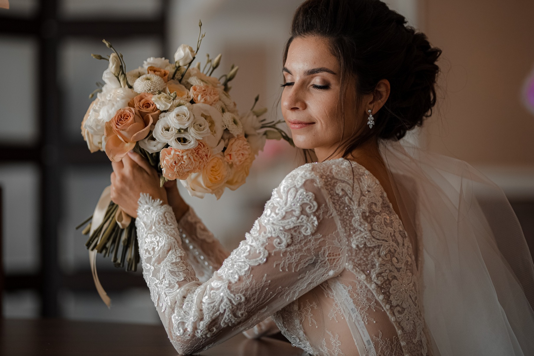 Panna młoda w koronkowej sukni z długim rękawem, trzymająca bukiet z jasnych róż i białych kwiatów, z zamkniętymi oczami i delikatnym uśmiechem, w miękkim, naturalnym świetle.