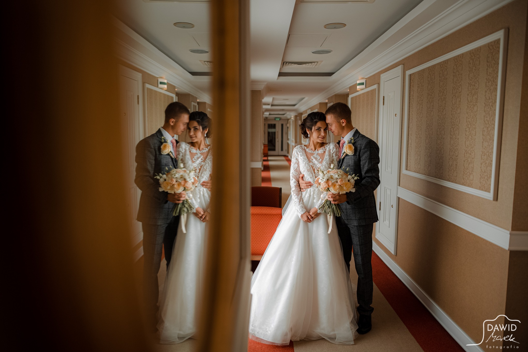 Elegancka para młoda w hotelowym korytarzu pozuje do zdjęcia, odbijając się w lustrze. Pan młody w kraciastej marynarce, panna młoda w koronkowej sukni trzyma bukiet.