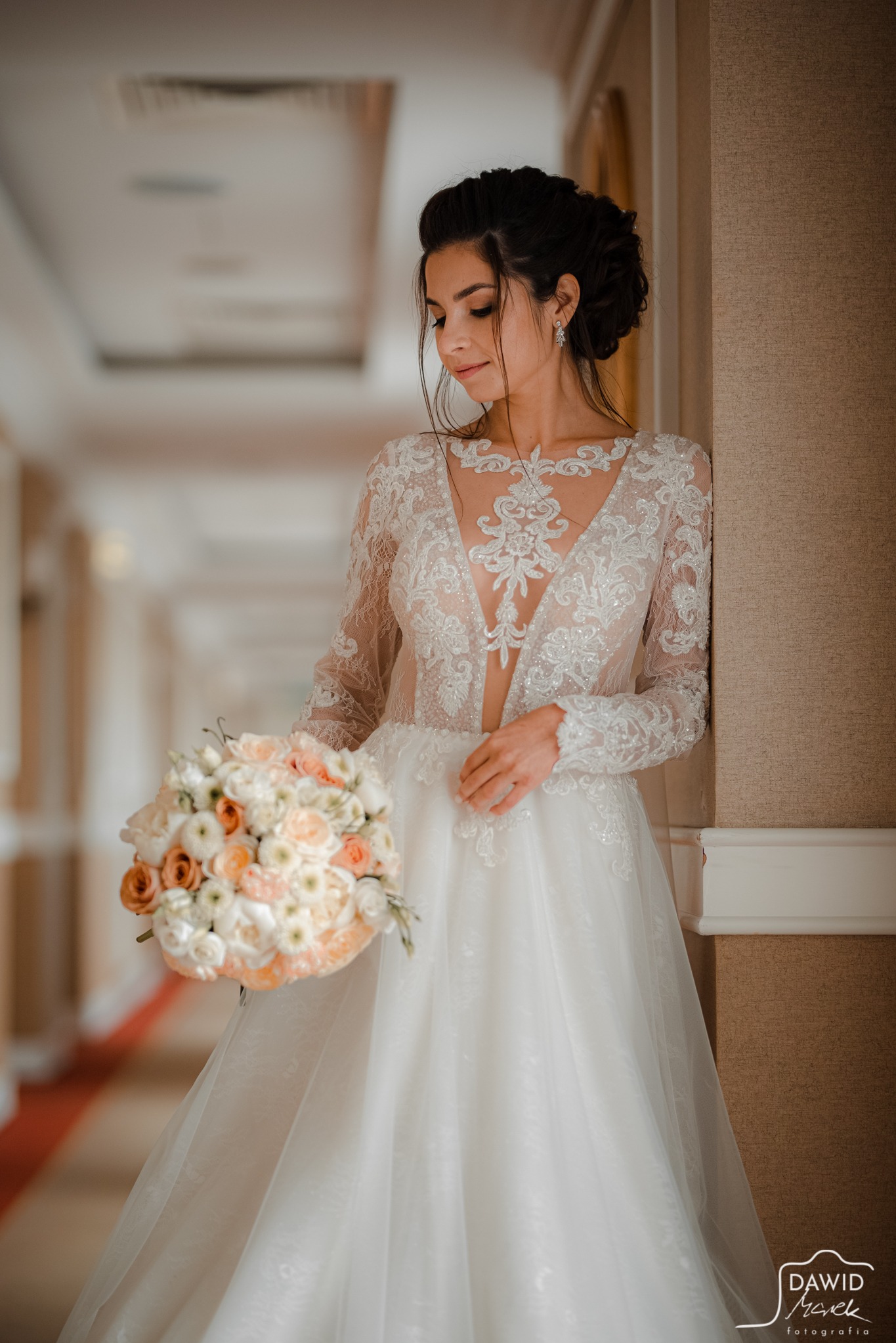 Panna młoda w koronkowej sukni z bukietem w pastelowych barwach, oparta o kolumnę w eleganckim wnętrzu. Subtelne światło podkreśla detale sukni.