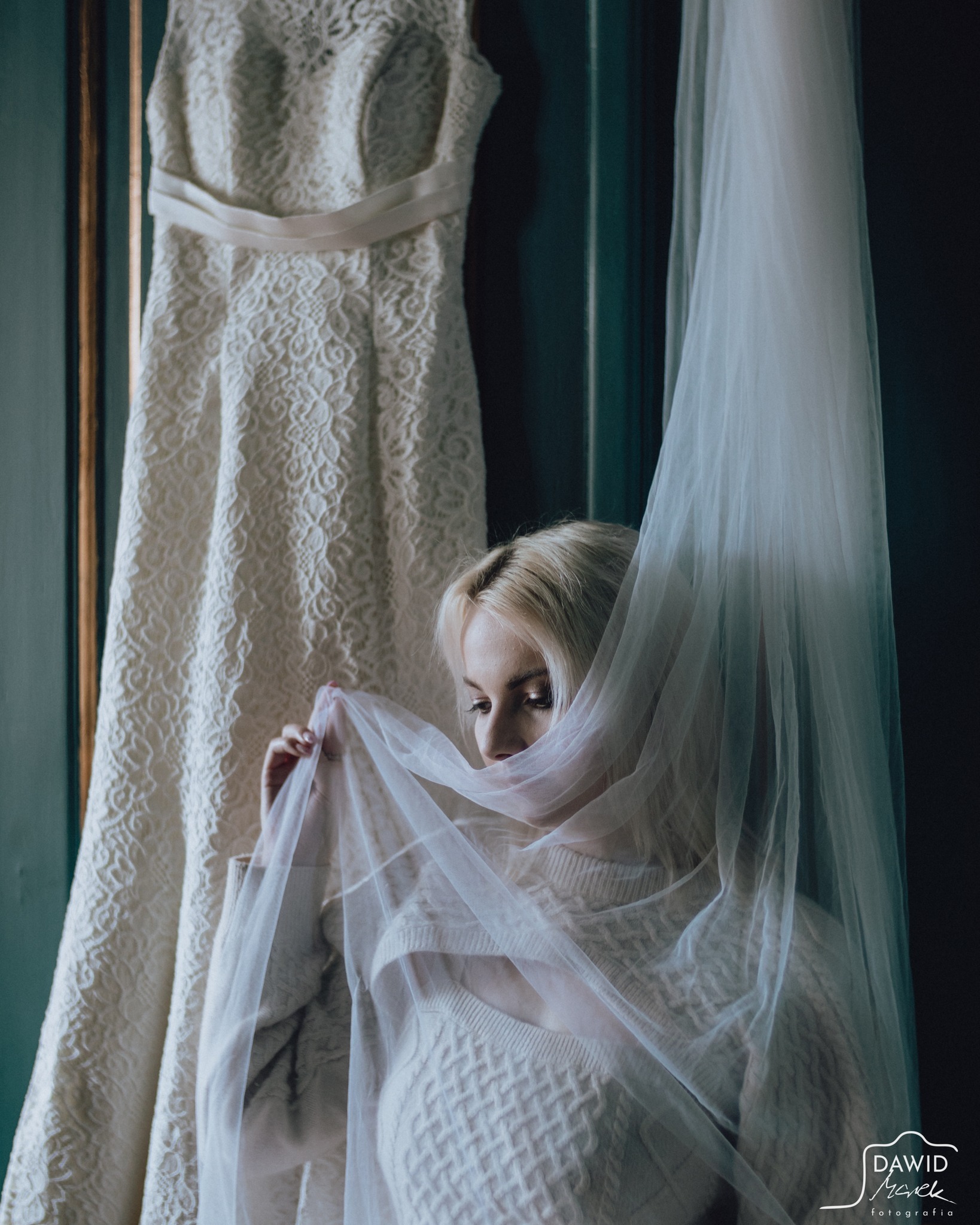 Panna młoda z blond włosami, częściowo zasłonięta woalką, pozuje obok sukni ślubnej z koronkowym wzorem, wiszącej na ciemnozielonym tle. Romantyczna atmosfera.