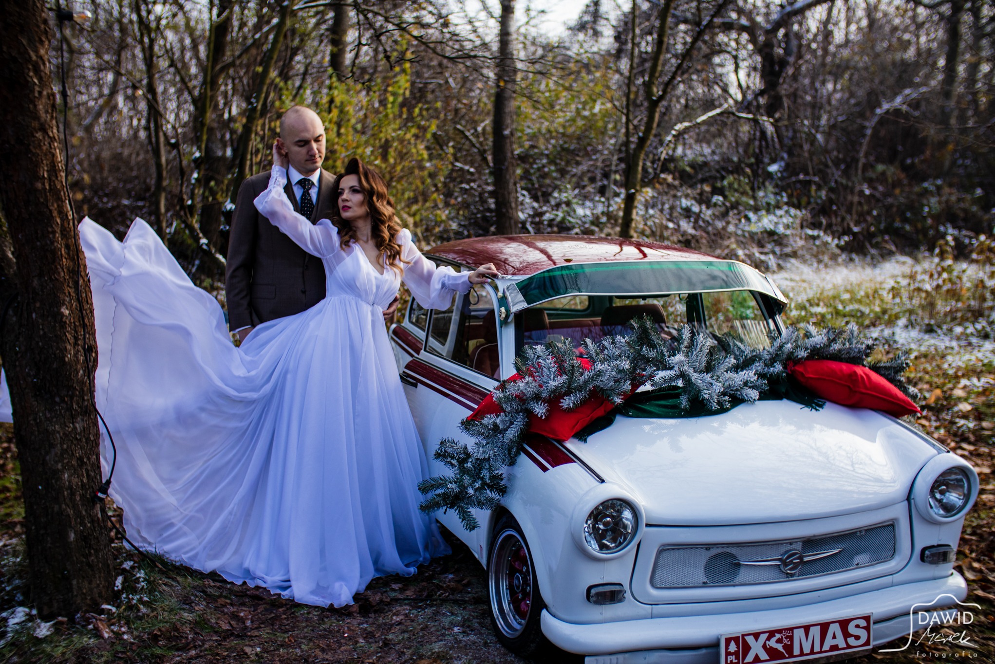 Para młoda w plenerze pozuje przy białym, udekorowanym na święta Trabancie. Kobieta w zwiewnej sukni, mężczyzna w garniturze. Zimowa sceneria, drzewa w tle.