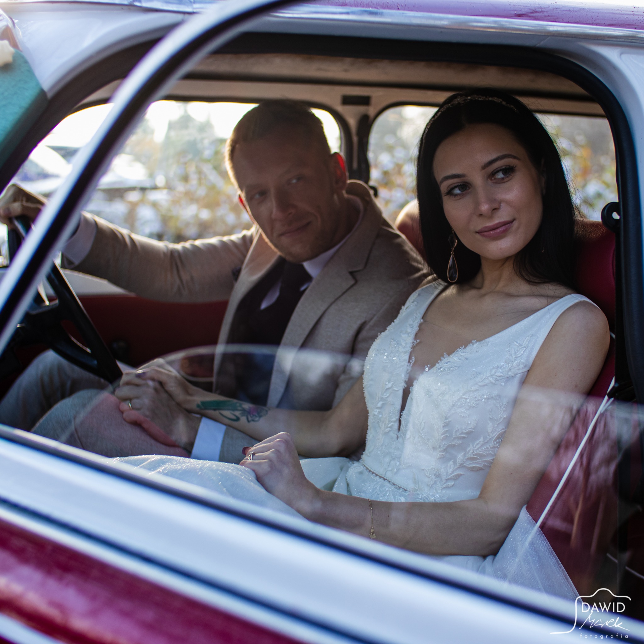 Para młoda siedzi w czerwono-białym samochodzie retro. Pan młody trzyma kierownicę, a panna młoda spogląda w bok. Ujęcie przez szybę, widoczny tatuaż na ręce pana młodego.