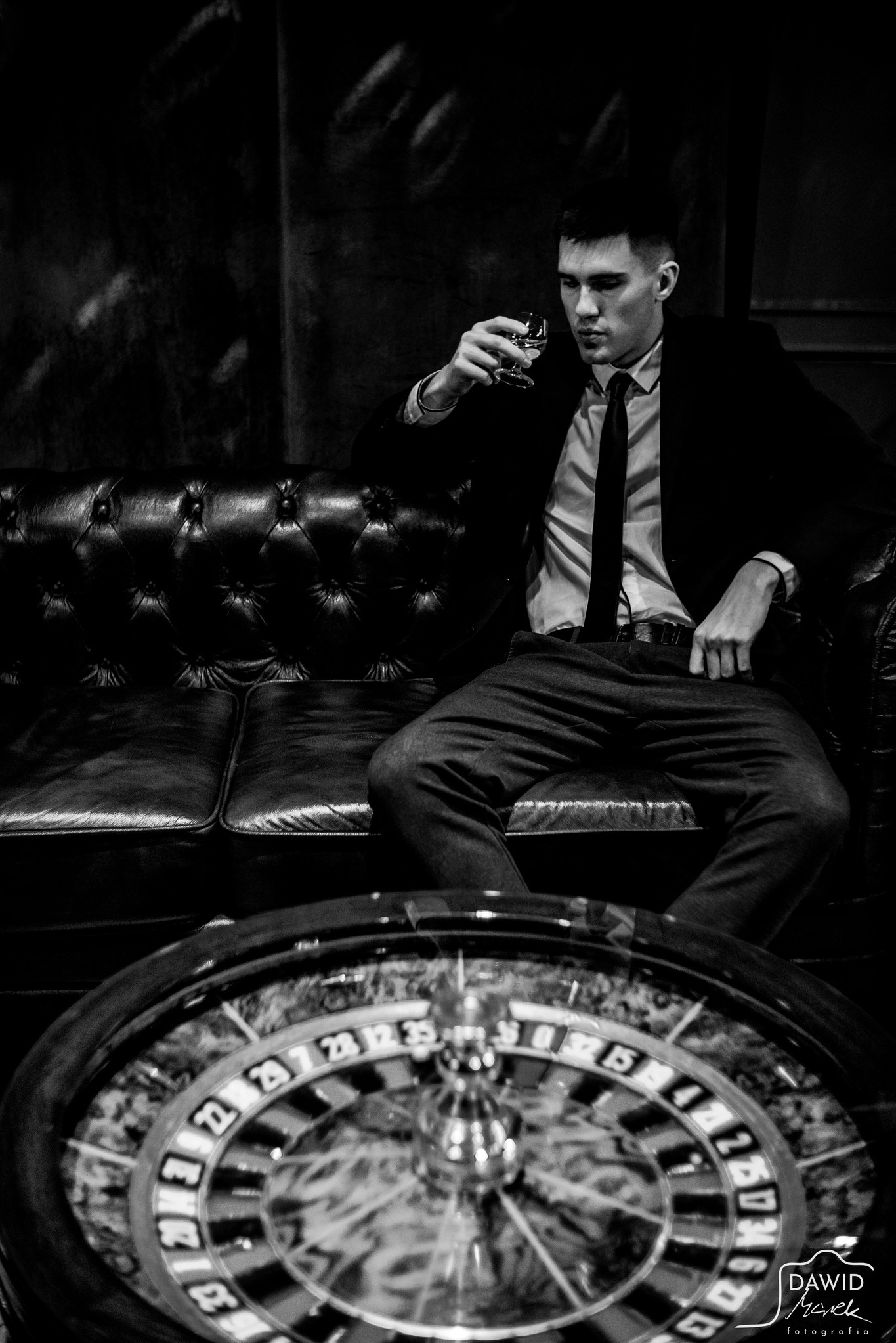 Czarno-białe zdjęcie mężczyzny w garniturze, siedzącego na skórzanej sofie z kieliszkiem w dłoni, obok ruletki. Klimat retro, elegancki styl.