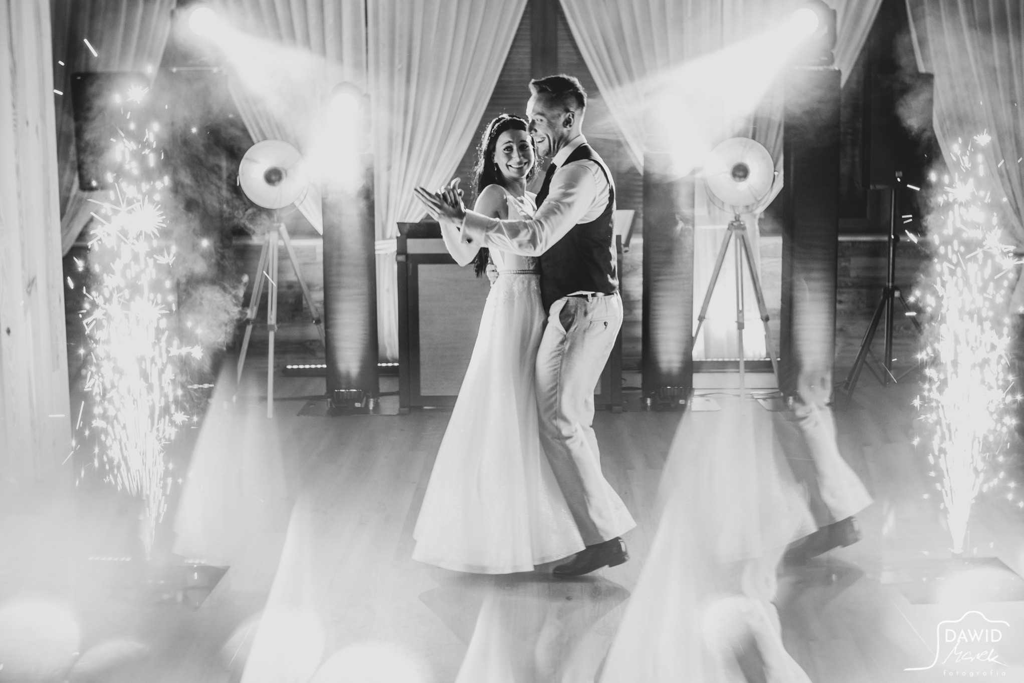 Czarno-białe zdjęcie uśmiechniętej pary młodej podczas pierwszego tańca, z iskrami fontann na parkiecie i reflektorami w tle, uchwycone z niskiego kąta.